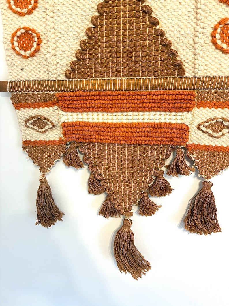 May include: Woven wall hanging with a geometric design in cream, brown, and orange. The textile features a variety of textures, including woven sections, tassels, and embroidered details. The wall hanging is mounted on a wooden dowel.