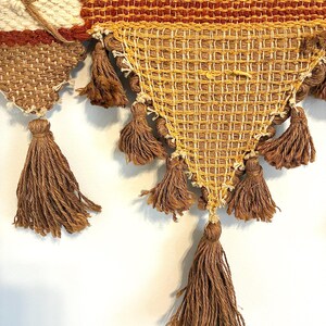 May include: Close-up of a woven wall hanging with a geometric design. The piece features a combination of cream, brown, and gold tones. Brown tassels hang from the bottom edge, adding texture and visual interest to the textile.