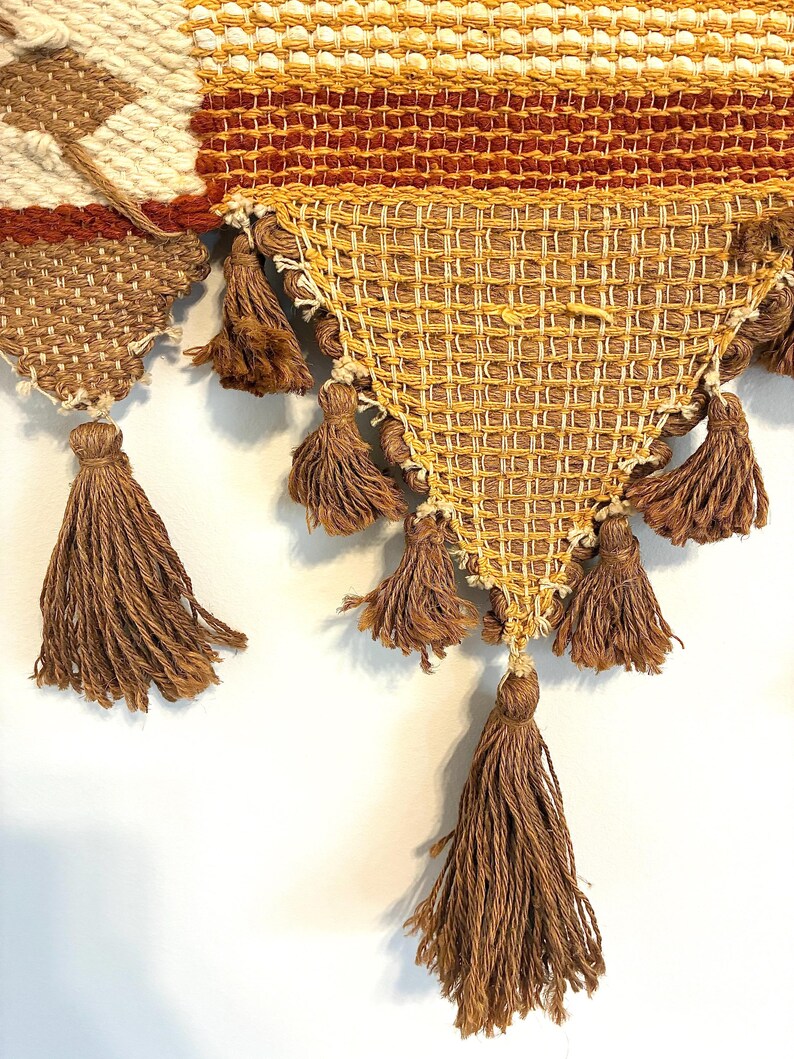 May include: Close-up of a woven wall hanging with a geometric design. The piece features a combination of cream, brown, and gold tones. Brown tassels hang from the bottom edge, adding texture and visual interest to the textile.