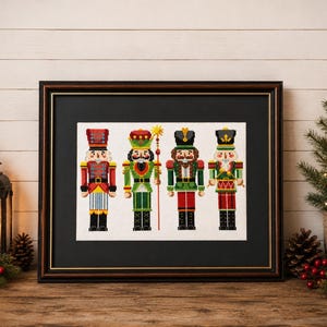 May include: A framed cross-stitch embroidery featuring four nutcracker figures in red, green, and gold uniforms. The figures are arranged side-by-side, each wearing a distinct hat and holding a staff or drum, displayed within a dark wooden frame.