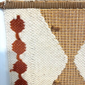 May include: Woven wall hanging with a geometric design. The textile features a cream-colored background with a diamond pattern in shades of brown and orange. A wooden rod and rope are at the top for hanging.