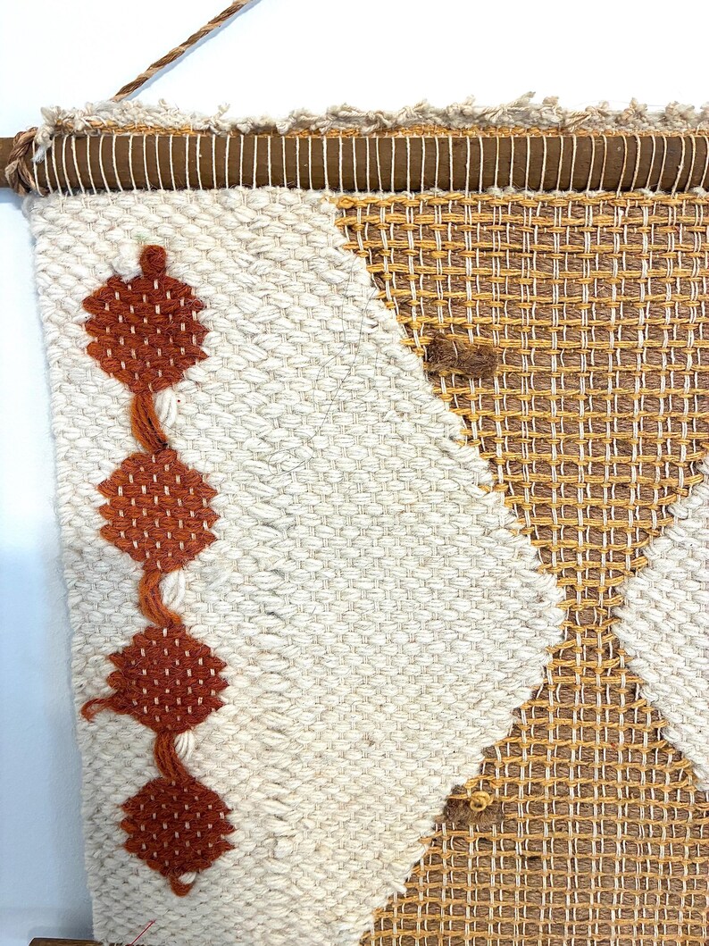 May include: Woven wall hanging with a geometric design. The textile features a cream-colored background with a diamond pattern in shades of brown and orange. A wooden rod and rope are at the top for hanging.