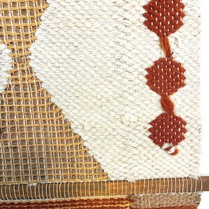 May include: A woven wall hanging with a geometric design. The piece features a combination of cream, brown, and rust-colored yarns. The design includes diamond and rectangular shapes, with a wooden dowel at the bottom for hanging. The overall texture is thick and tactile.