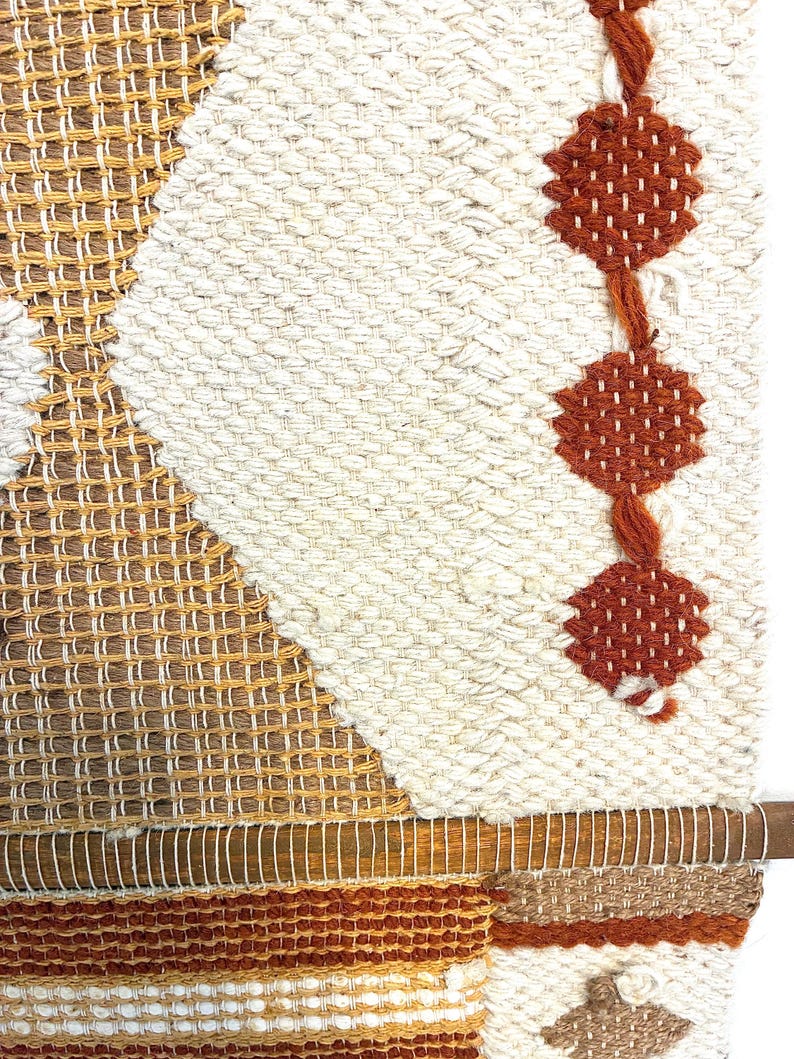 May include: A woven wall hanging with a geometric design. The piece features a combination of cream, brown, and rust-colored yarns. The design includes diamond and rectangular shapes, with a wooden dowel at the bottom for hanging. The overall texture is thick and tactile.