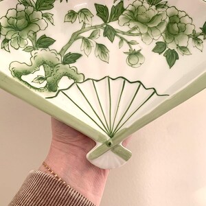 Op de afbeelding: Witte, waaiervormige decoratieve schaal met een groen bloemenontwerp. De schaal heeft een handgeschilderd patroon van bloemen en bladeren. De randen zijn groen omlijnd en de schaal wordt door een hand vastgehouden.