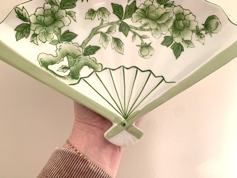 Op de afbeelding: Witte, waaiervormige decoratieve schaal met een groen bloemenontwerp. De schaal heeft een handgeschilderd patroon van bloemen en bladeren. De randen zijn groen omlijnd en de schaal wordt door een hand vastgehouden.