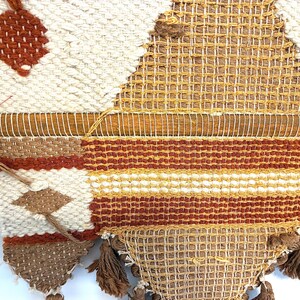 May include: A woven wall hanging with a geometric design. The piece features a wooden dowel at the top and is made with cream, brown, and orange yarns. The design includes diamond and striped patterns, with tassels at the bottom.