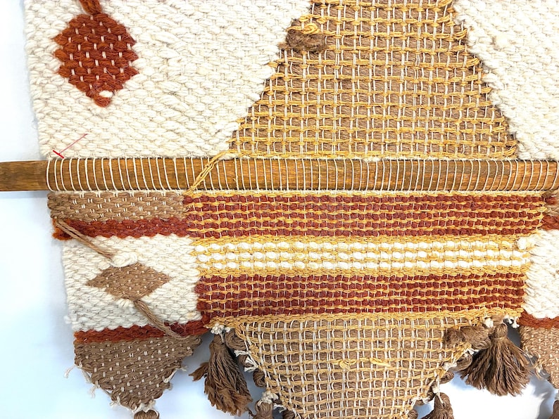 May include: A woven wall hanging with a geometric design. The piece features a wooden dowel at the top and is made with cream, brown, and orange yarns. The design includes diamond and striped patterns, with tassels at the bottom.
