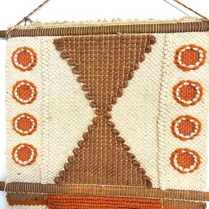 May include: Woven wall hanging with a geometric design. The piece features a cream-colored background with brown and orange accents. The design includes a central hourglass shape and circular patterns. The wall hanging is suspended from a wooden rod.