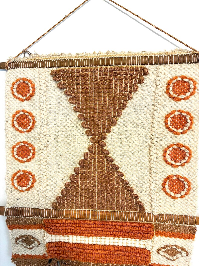May include: Woven wall hanging with a geometric design. The piece features a cream-colored background with brown and orange accents. The design includes a central hourglass shape and circular patterns. The wall hanging is suspended from a wooden rod.