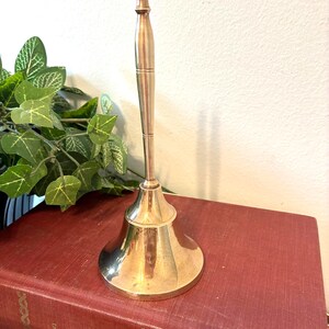 Op de afbeelding: Een glanzende messing bel met een lange handgreep en een uitlopende basis. De bel staat op een vintage rood boek. De achtergrond omvat groene planten en een witte muur.