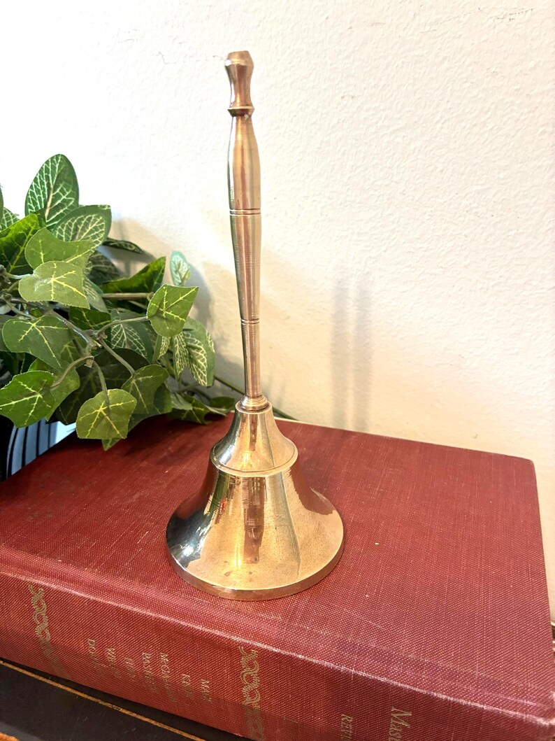 Op de afbeelding: Een glanzende messing bel met een lange handgreep en een uitlopende basis. De bel staat op een vintage rood boek. De achtergrond omvat groene planten en een witte muur.