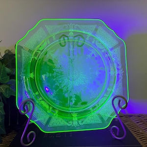 May include: A square-shaped, translucent green glass plate featuring an intricate, etched floral and geometric pattern. The plate glows with a bright neon green hue under ultraviolet light, highlighting the detailed decorative border and center.