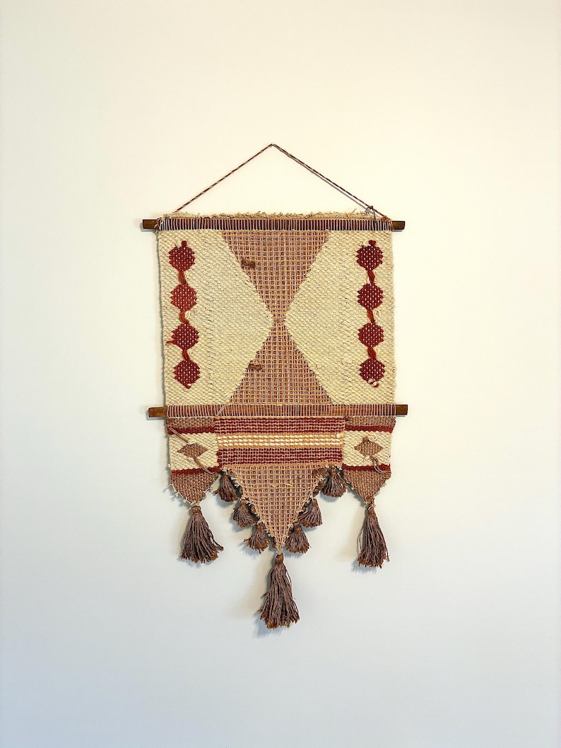 May include: Woven wall hanging with a geometric design. The tapestry features a cream-colored background with a diamond pattern in shades of brown and red. Brown tassels hang from the bottom, and the piece is suspended from a wooden rod.