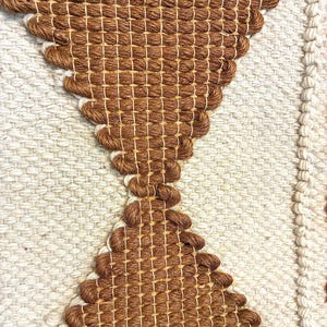 May include: Close-up of a woven textile with a geometric design. The piece features a central brown hourglass shape against a cream-colored background. The texture is visible, with the weave creating a tactile surface.