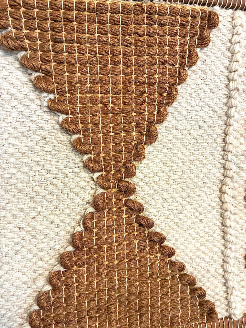 May include: Close-up of a woven textile with a geometric design. The piece features a central brown hourglass shape against a cream-colored background. The texture is visible, with the weave creating a tactile surface.