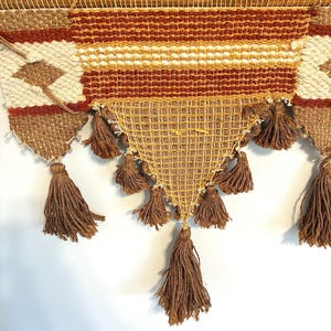 May include: A woven wall hanging with a geometric design in shades of brown, cream, and orange. The piece features a triangular bottom section with multiple brown tassels. The top section has horizontal stripes and diamond patterns.