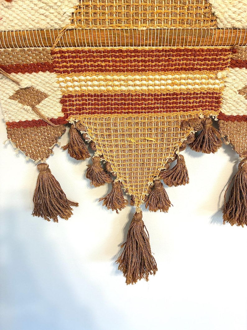 May include: A woven wall hanging with a geometric design in shades of brown, cream, and orange. The piece features a triangular bottom section with multiple brown tassels. The top section has horizontal stripes and diamond patterns.