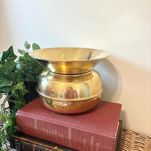 May include: A vintage brass decorative bowl with a wide, shallow top and a rounded base. The bowl is sitting on top of two antique books with red and brown covers. The books are stacked on a woven wicker surface.