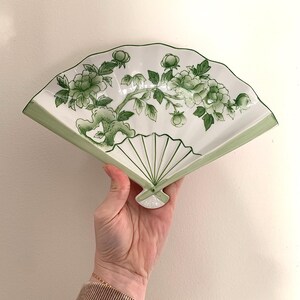 Op de afbeelding: Een witte, keramische, waaiervormige decoratieve plaat met een groen bloemmotief. De plaat heeft een groene rand en een gedetailleerde illustratie van bloemen en bladeren. De plaat wordt in een hand gehouden.