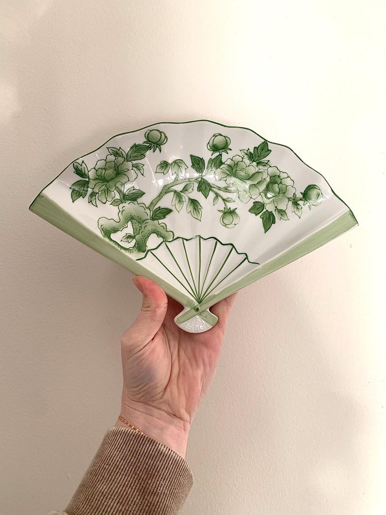 Op de afbeelding: Een witte, keramische, waaiervormige decoratieve plaat met een groen bloemmotief. De plaat heeft een groene rand en een gedetailleerde illustratie van bloemen en bladeren. De plaat wordt in een hand gehouden.