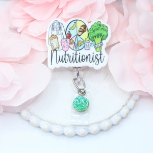 May include: A white badge reel with a colorful illustration of a food pyramid and the word "Nutritionist". The reel has a green glitter charm attached.
