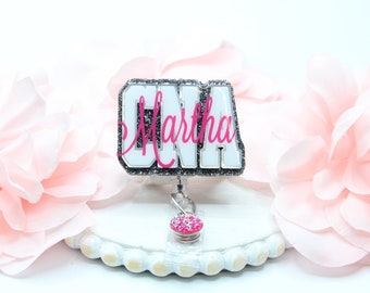 May include: A white and silver badge holder with the word "CNA" in large white letters and the name "Martha" in pink letters. The badge holder has a pink and silver retractable badge reel.