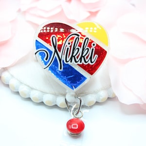 May include: A red, white, and blue heart-shaped badge holder with the name "Nikki" written in black. The badge holder is attached to a retractable badge reel with a red button.