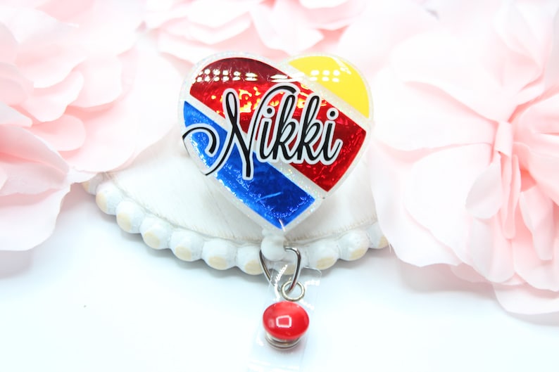 May include: A red, white, and blue heart-shaped badge holder with the name "Nikki" written in black. The badge holder is attached to a retractable badge reel with a red button.