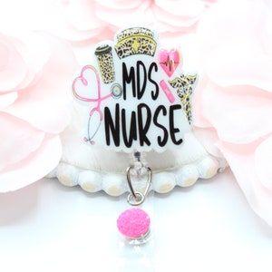 May include: A white MDS Nurse badge reel with a pink glitter accent. The badge features a coffee cup, nurse's cap, stethoscope, and heart with a heartbeat graphic. The text "MDS NURSE" is in black.