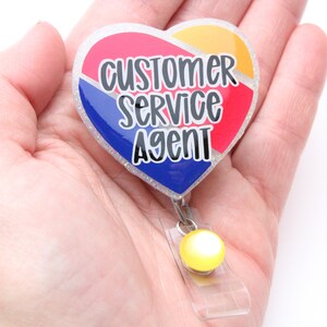 May include: A heart-shaped badge holder with a glitter finish. The badge features a red, white, and blue design with the text "Customer Service Agent".