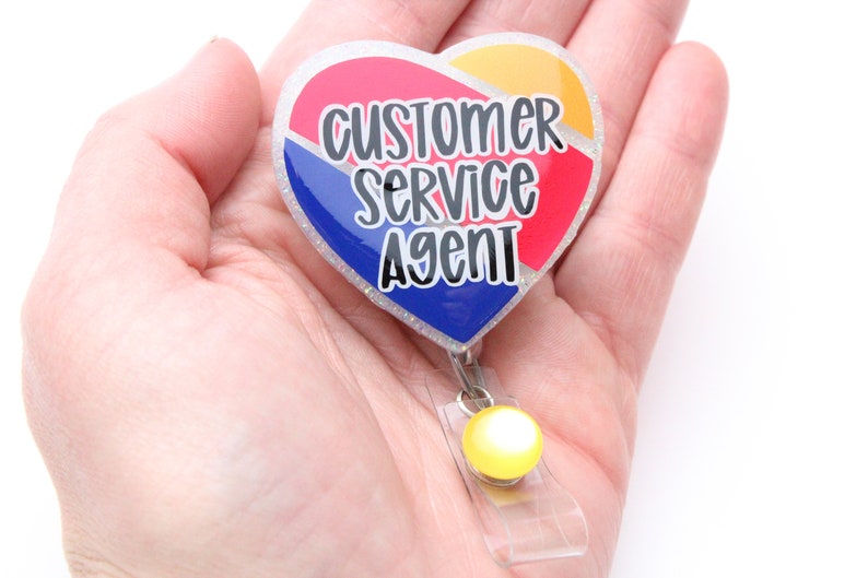 May include: A heart-shaped badge holder with a glitter finish. The badge features a red, white, and blue design with the text "Customer Service Agent".