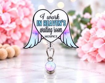 Könnte beinhalten: Ein herzförmiger Ausweishalter mit Engelsflügeln in einem blau-rosa Farbverlauf. Der Text lautet I work IN HEAVEN'S waiting room HOSPICE. Eine kleine, runde, irisierende Perle hängt an einem silbernen Metallring an der transparenten Kunststoffbasis.