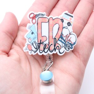 May include: A white and pink badge reel with the text "ER Tech" and medical illustrations. The reel has a blue button and a clear plastic cord.