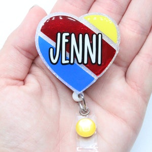 May include: A heart-shaped badge holder with a red, blue, and yellow design. The name "JENNI" is written in white letters on the badge holder. The badge holder has a yellow retractable badge reel.