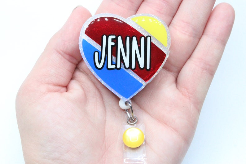 May include: A heart-shaped badge holder with a red, blue, and yellow design. The name "JENNI" is written in white letters on the badge holder. The badge holder has a yellow retractable badge reel.