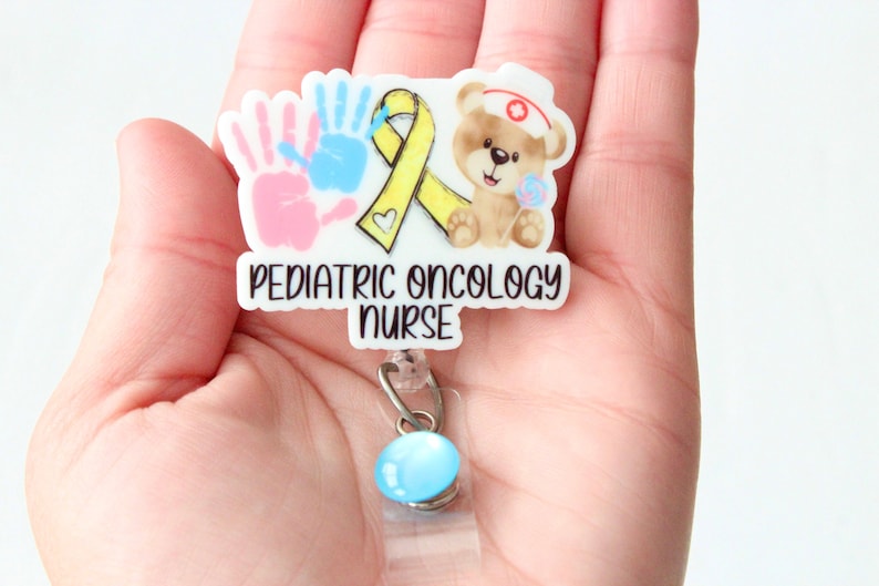 May include: White badge featuring a yellow ribbon, two handprints, and a teddy bear wearing a nurse's hat. The badge reads "Pediatric Oncology Nurse".