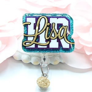 May include: A teal blue and purple glitter badge holder with the name "Lisa" in gold lettering. The badge holder has a gold glitter ball charm attached to it.