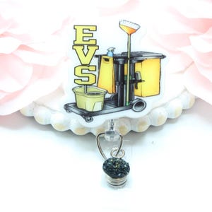 May include: A white badge reel with a black and yellow cleaning cart and the letters EVS. The badge reel has a black crystal charm.