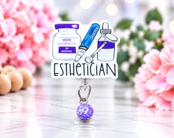May include: A badge reel featuring a white acrylic topper with a graphic illustration of a Botox vial, a blue Skin Pen, and a medical tool. The text ESTHETICIAN is printed in black below the items. A small purple bead hangs from the metal clip.