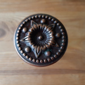 May include: Close-up of a decorative bronze cabinet knob. The knob features a central floral design surrounded by a ring of small, raised dots. The bronze has a slightly aged appearance, with hints of green patina.