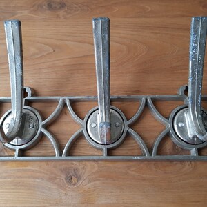 Puede incluir: Un perchero de pared de metal vintage con tres ganchos. Los ganchos son curvos y están unidos a un marco decorativo con un diseño geométrico. El metal tiene un aspecto deslustrado y envejecido, lo que sugiere que es antiguo.