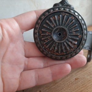 May include: Close-up of a decorative, antique-style cabinet knob. The knob is round, with a dark, textured finish and a central design of radiating lines. The edges are detailed with small, raised dots. The metal has a distressed, aged appearance.