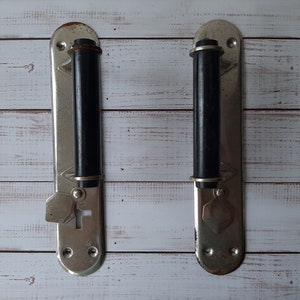 May include: Two vintage door handles with a silver finish and black cylindrical grips. Each handle has an elongated backplate with decorative elements and keyhole details, suggesting an antique style.