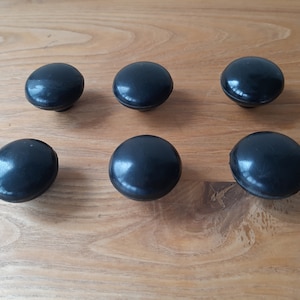 May include: Six black, round cabinet knobs with a glossy finish are arranged on a light brown wooden surface. The knobs have a simple, classic design, suitable for various furniture styles. Each knob is approximately 1.5 inches in diameter.