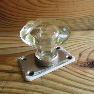 May include: Clear glass door knob with a decorative, rounded top. The knob is mounted on a rectangular metal plate with a distressed white finish. The background is a wooden surface.