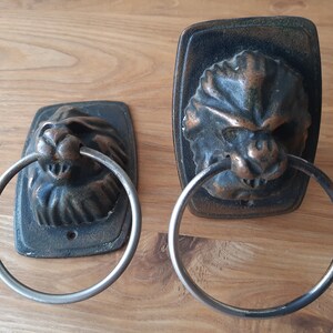 May include: Two antique bronze lion head door knockers with silver-colored rings. The lion heads are detailed with textured manes and facial features. The knockers are mounted on rectangular backplates.