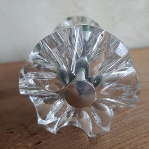 May include: A clear, faceted glass bottle stopper with a silver-colored metal base. The stopper has a starburst design and is likely for a wine or liquor bottle. The glass is transparent, allowing light to pass through.