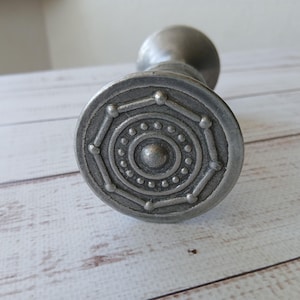 Puede incluir: Un tapón de botella plateado con un diseño circular decorativo. El tapón presenta una serie de círculos concéntricos y un punto central en relieve, con un vástago que se extiende hacia arriba. El diseño es detallado y texturizado, lo que sugiere un estilo vintage o antiguo.