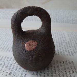 May include: A dark brown, textured cast iron weight with a rounded handle and a circular copper-colored inlay on the front. The inlay features a small stamped crown symbol above the number 114, clearly visible against the dark metal surface.